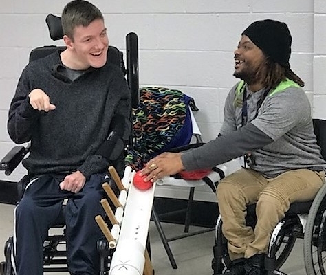 Coach in wheelchair assisting athlete in powerwheelchair use ramp to play boccia. Both are smiling and laughing