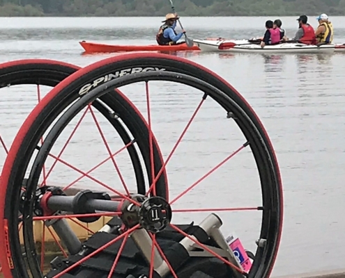 4 people in kayaks in the distance on the lake. In the foreground, is a wheelchair on the dock