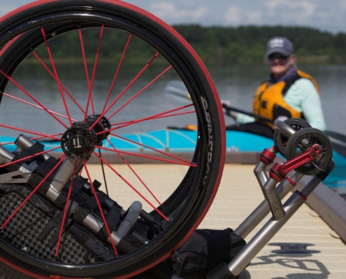 picture of wheelchair in forefront on dock. Blurred in the background in a kayak on the water is a woman wearing a hat- Ashley Thomas.