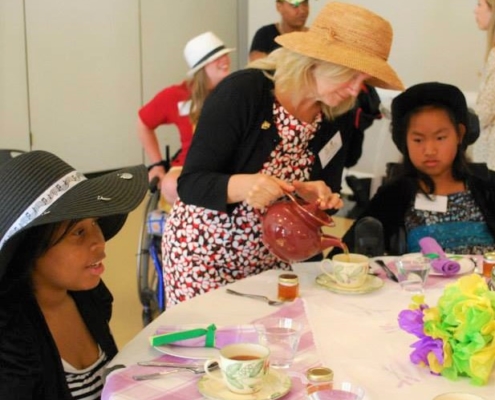 Woman in fancy hat pours tea at a decorated tea partyfor young girl using wheelchair at GiGe event in 2014at