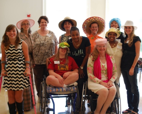 group of women and girls some using wheelchairs and forearm crutches smile in group wearing fancy hats at GiGe event in 2014